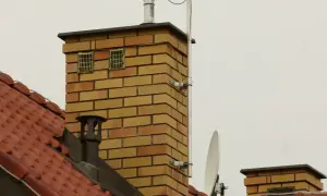 Na dachu ceglanego komina widać dwa kratkowane otwory, które mogą służyć jako odpowietrzenie kanalizacji. Obok anteny satelitarnej i innych elementów instalacji.