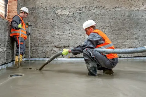 Dwóch budowlańców w kamizelkach odblaskowych wylewa pianobeton przez wąż na podłogę.