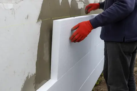 Pracownik w czerwonych rękawicach montuje płyty styropianowe na ścianie domu, tworząc izolację elewacji.