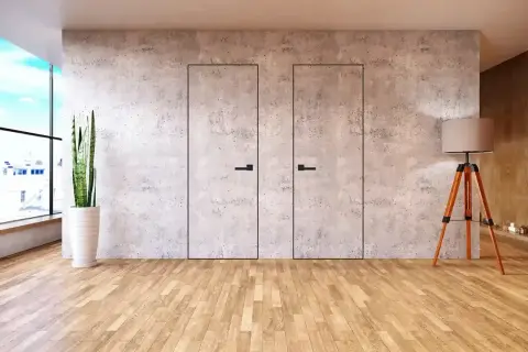 Dwa minimalistyczne, **drzwi ukryte w ścianie** z betonu architektonicznego, z czarnymi uchwytami. Obok stoi lampa na trójnogu.