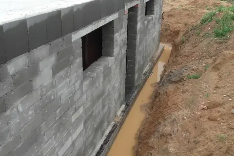 Budowa fundamentów z bloczków betonowych, obok wykop z wodą, który wymaga drenażu.
