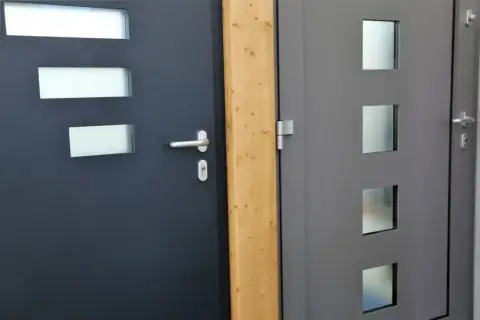 Dwie nowoczesne drzwi, jedna ciemnoszara z trzema prostokątnymi przeszkleniami, druga szara z trzema kwadratowymi, obok drewnianego słupka. Dowiedz się, jak wygłuszyć drzwi.