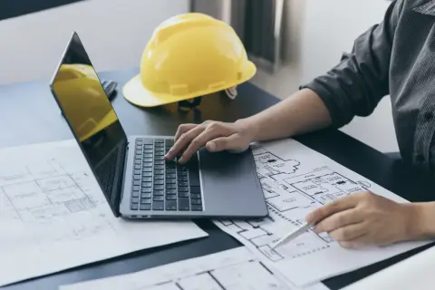 Architekt analizuje plany, obliczając powierzchnię użytkową. Na biurku laptop, żółty kask i rysunki techniczne.