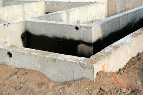 Surowe ściany fundamentowe z betonu, częściowo pokryte czarną izolacją przeciwwilgociową, otoczone ziemią.