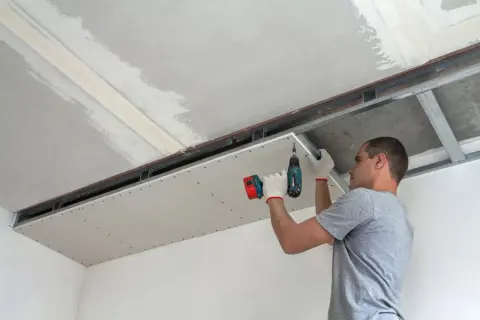 Mężczyzna montuje płyty regips do stelaża sufitowego, używając wkrętarki.