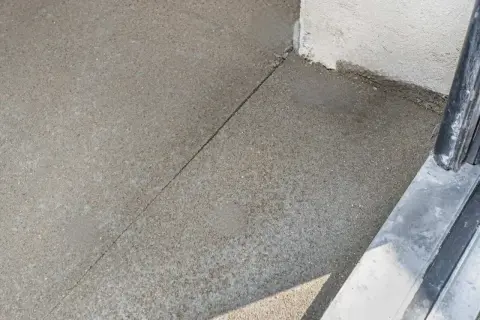Szara betonowa posadzka z widoczną linią dylatacji, biegnącą od narożnika budynku do metalowej balustrady.