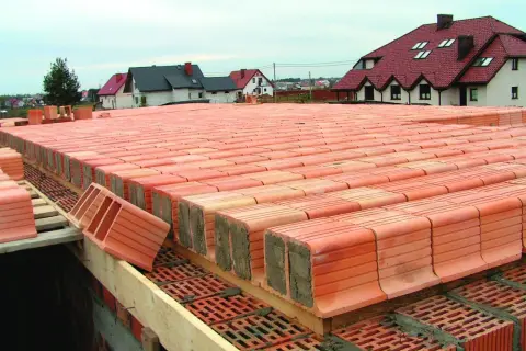 Strop ackermana w budowie, rzędy pustaków ceramicznych czekają na betonowanie. W tle domy z czerwonymi dachami.