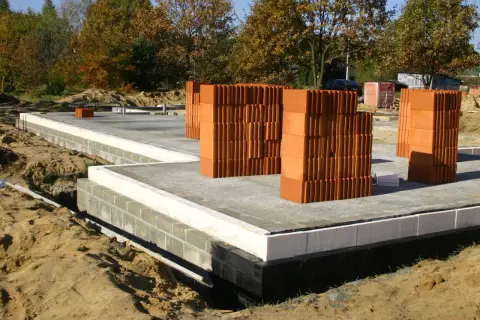 Stosy cegieł, gotowe do budowy domu. Fundament z bloczków i izolacji, piasek wokół. Chudziak czeka na swoją kolej.