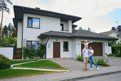 Szczęśliwa rodzina przed nowoczesną zabudową zagrodową. Dziewczynka siedzi na ramionach ojca, rozkładając ręce.