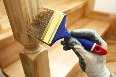 Ręka w rękawicy maluje pędzlem drewniany słupek schodów.