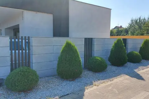 Nowoczesny betonowy płot z ozdobnymi krzewami i bramą.