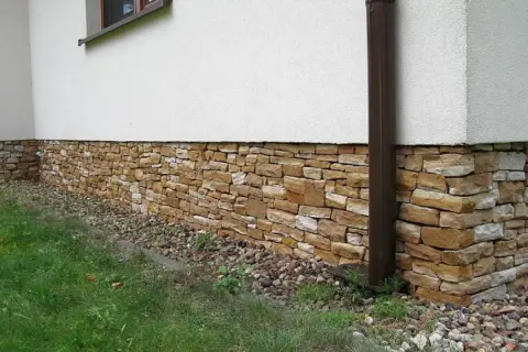 Kamienny cokół na elewacji domu, wykonany z nieregularnych, piaskowych kamieni, otoczony żwirem i zieloną trawą.