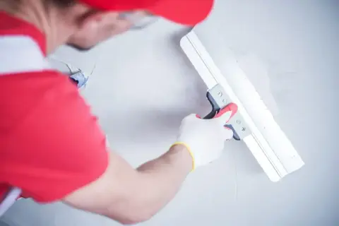 Gipsar w czerwonej czapce i rękawiczkach wygładza ścianę gładzią za pomocą szerokiej pacy.