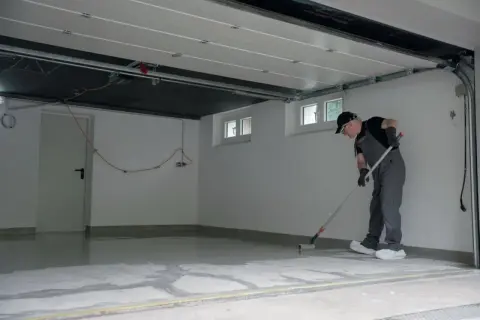 Mężczyzna w kombinezonie maluje wałkiem podłogę w garażu. Nowa, lśniąca żywica w garażu odmieni jego wygląd.