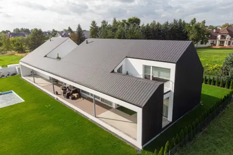 Nowoczesny dom z szarą blachą na rąbek, otoczony zielonym ogrodem i basenem.