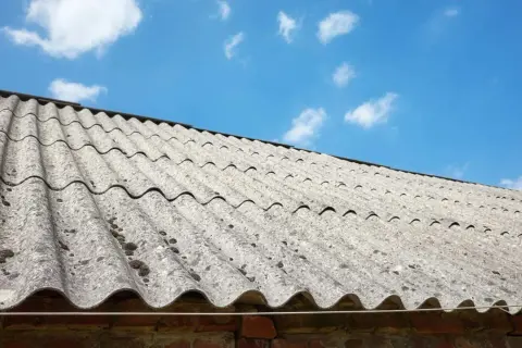 Stary dach pokryty eternitem, materiałem zawierającym azbest, pod błękitnym niebem z chmurami.