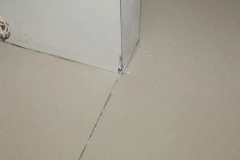 Uszkodzona dylatacja posadzki w rogu pomieszczenia, widoczne pęknięcia i ubytki materiału.