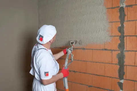 Pracownik nakłada tynk maszynowy na ścianę z pustaków ceramicznych. Sprawdź atrakcyjną tynki maszynowe cenę!