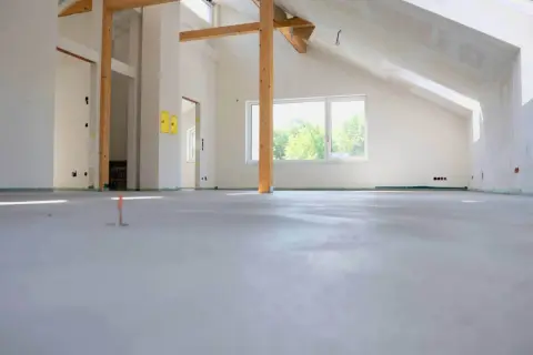 Nowe wnętrze z widoczną, świeżo wylaną wylewką samopoziomującą. Trwają prace budowlane, widoczne drewniane belki stropowe i okna z widokiem na zieleń.