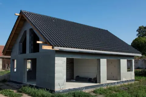 Dom w stanie surowym otwartym, z dachem pokrytym czarną dachówką. Widoczne ściany z bloczków, otwory okienne i drzwiowe.