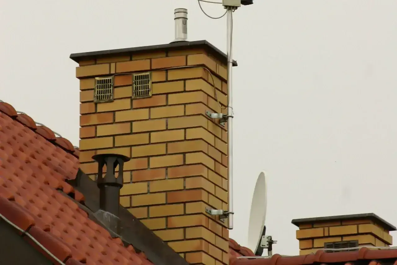 Na dachu ceglanego komina widać dwa kratkowane otwory, które mogą służyć jako odpowietrzenie kanalizacji. Obok anteny satelitarnej i innych elementów instalacji.